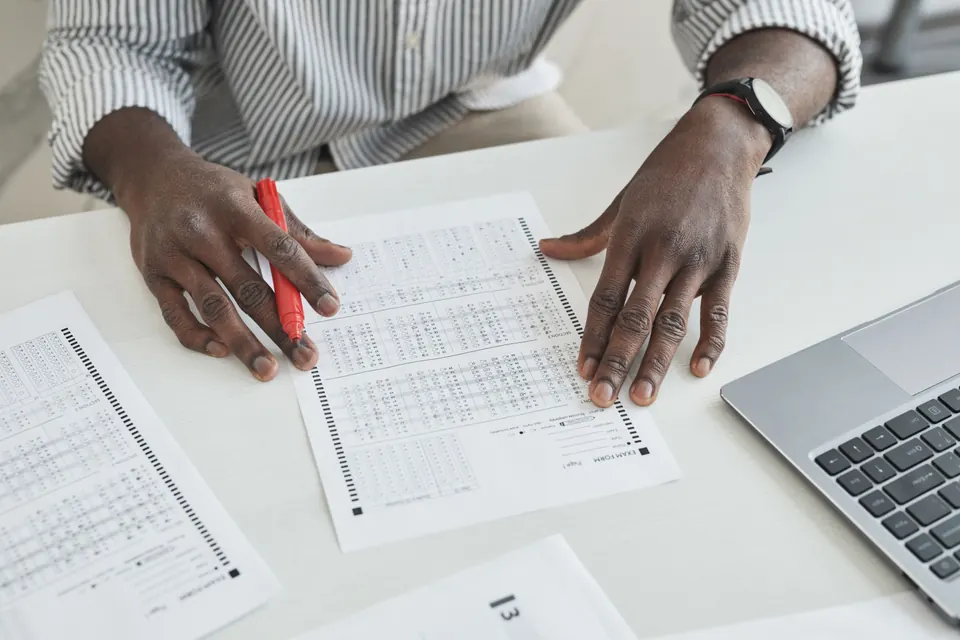 Two hands on a printed answer sheet, one holding a red marker, with more sheets and a laptop on a white desk.
