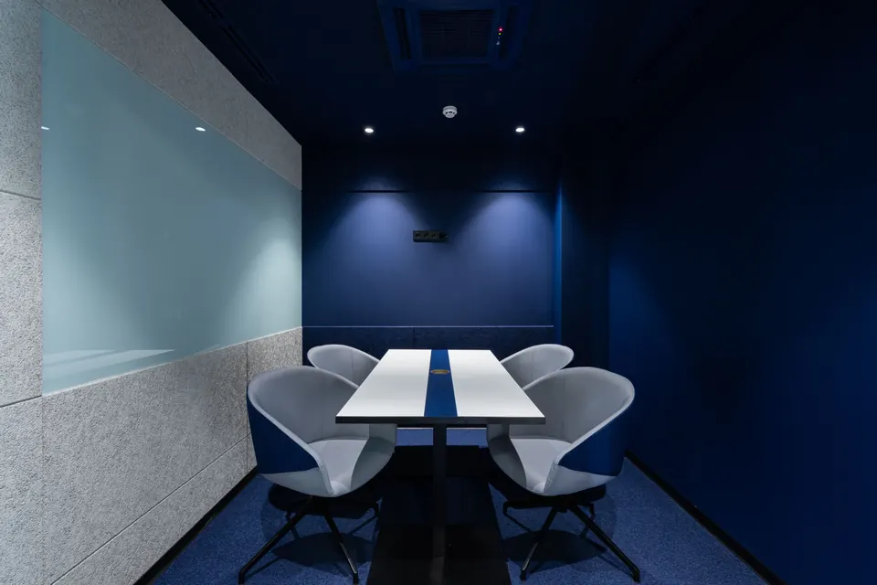 Petite salle de réunion fermée aux murs bleu nuit, table blanche à bande bleue et quatre fauteuils gris sous des spots