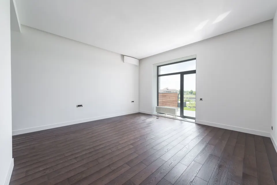 Pièce vide et lumineuse aux murs blancs, parquet foncé et porte-fenêtre vitrée donnant sur un jardin