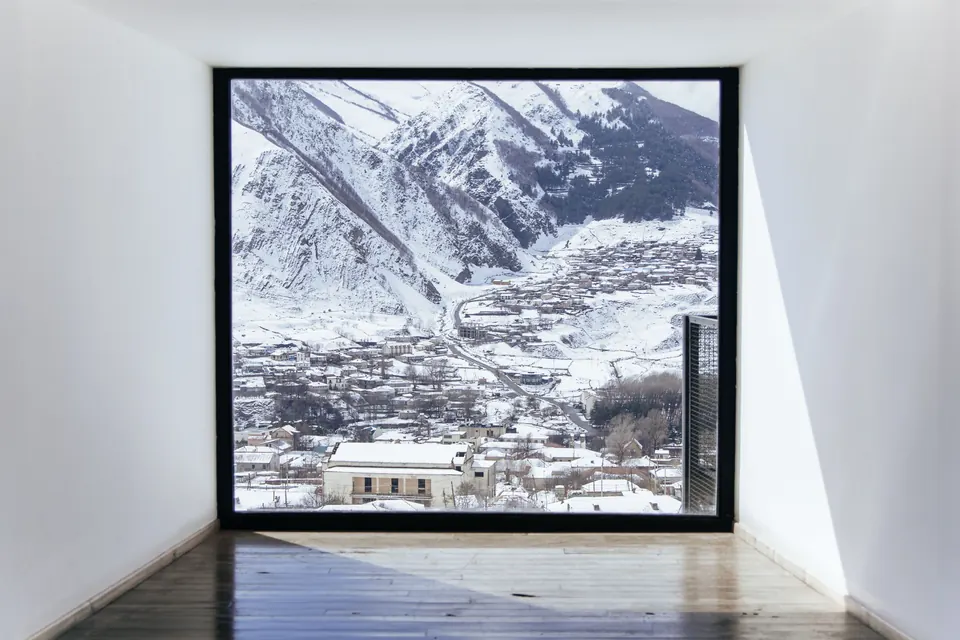 Bright empty room with a large square window opening onto a snow-covered mountain valley and a village below