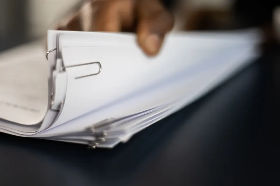 Eine Hand hebt die obersten Blätter eines dicken Stapels weißer Seiten an, die mit Büroklammern gehalten werden, auf dunklem Tisch