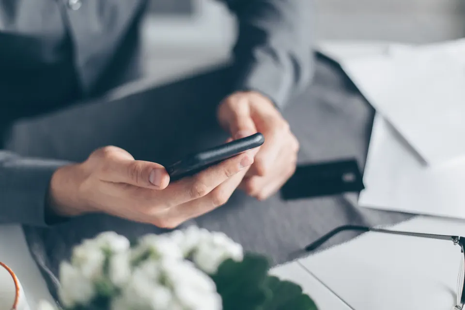 Two hands holding a black smartphone above a grey desk, with white papers, reading glasses and white flowers nearby