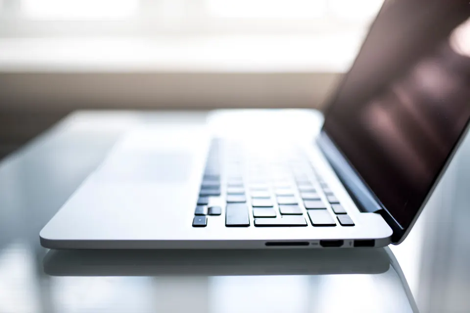 Open silver laptop seen from the side on a glossy white table, keyboard in focus, screen dark, bright window behind