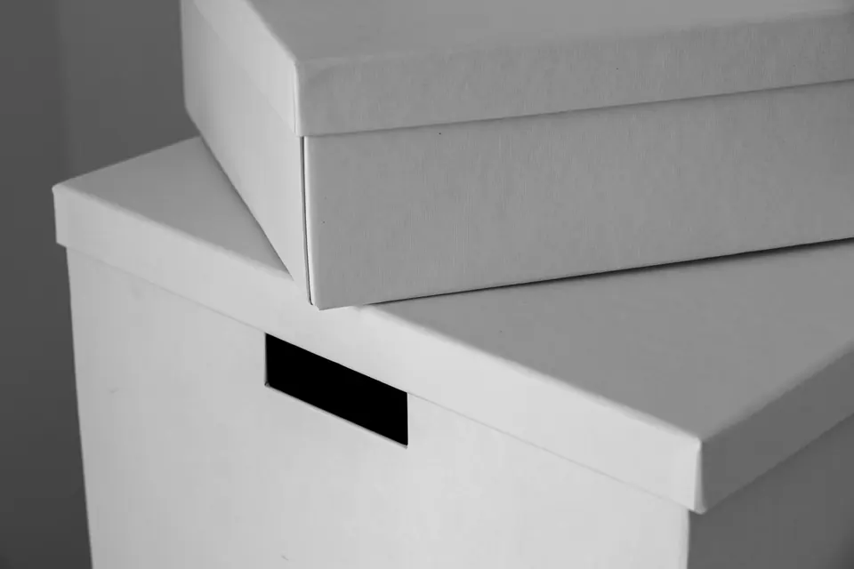 Deux boîtes d'archives blanches empilées dans une lumière grise, celle du bas percée d'une poignée rectangulaire noire